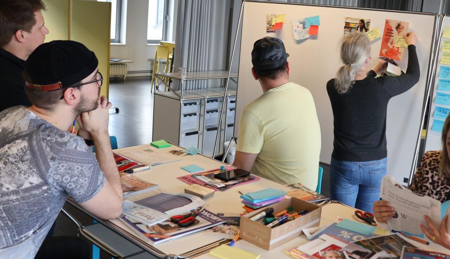 Auf dem Bild arbeiten Menschen zusammen. Sie sitzen am Tisch oder kleben Notizzettel an eine Magnetwand