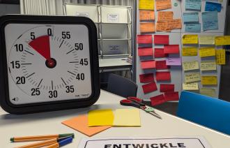 Auf dem Bild ist ein Workshopwecker abgebildet, der auf einem Tisch mit Stiften und PostIts steht. Im Hintergrund ist eine Magnetwand zu sehen, die voller Notizen ist.