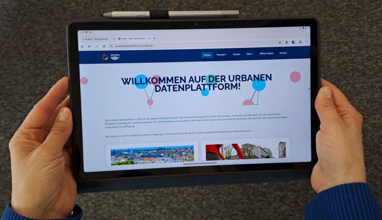 Zu sehen sind zwei Haende, die ein Tablet halten. Auf dem Tablet wurde die Webseite der Urbanen Datenplattform Potsdam aufgerufen.