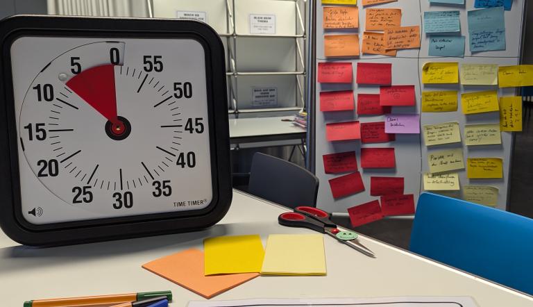 Auf dem Bild ist ein Workshopwecker abgebildet, der auf einem Tisch mit Stiften und PostIts steht. Im Hintergrund ist eine Magnetwand zu sehen, die voller Notizen ist.
